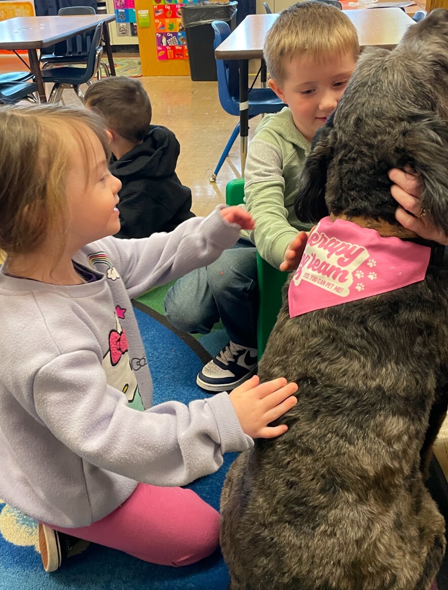 Two preschool children petting a dog who is wearing a pink kerchief.